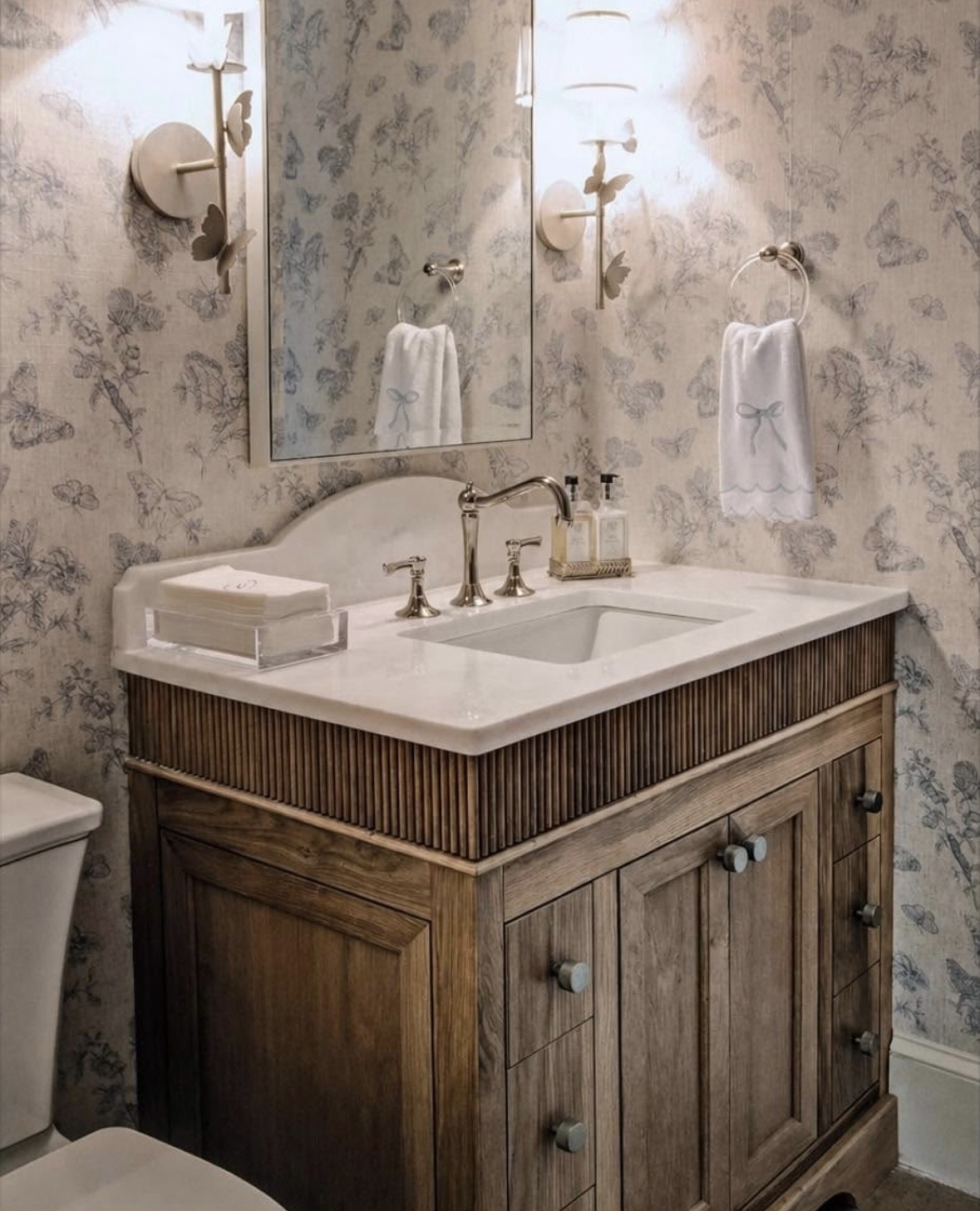 Fluted walnut vanity with marble countertop, polished faucet, and butterfly sconces